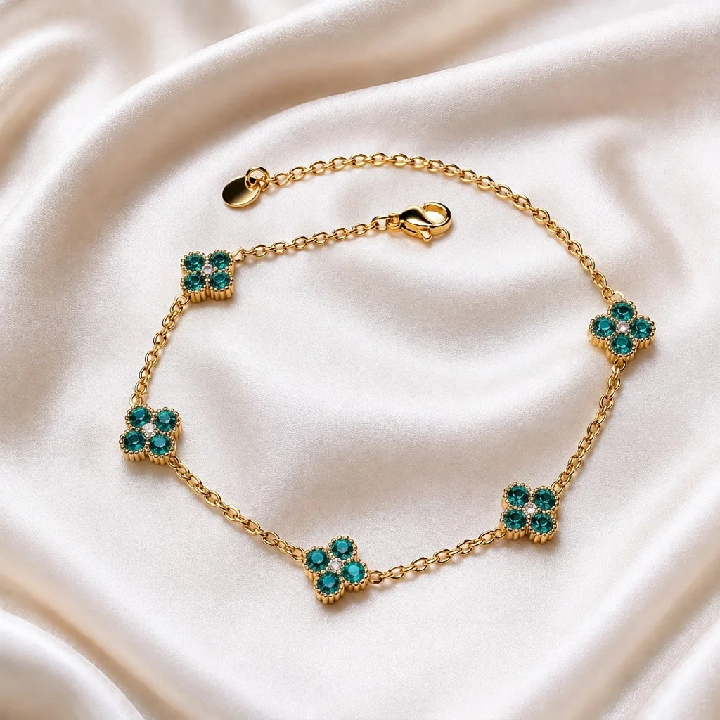 A gold chain bracelet with five floral clover motifs, each set with green and clear rhinestones, displayed on a draped cream satin fabric.