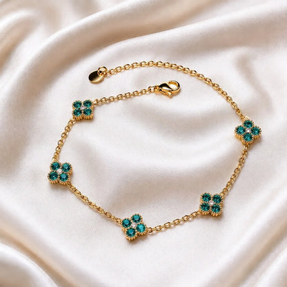 A gold chain bracelet with five floral clover motifs, each set with green and clear rhinestones, displayed on a draped cream satin fabric.