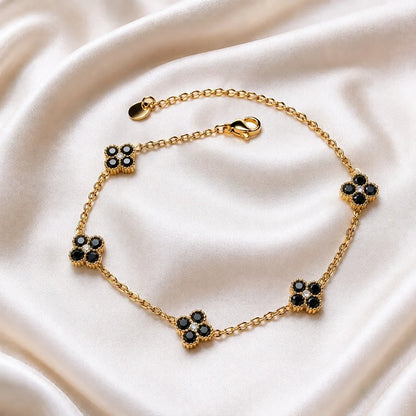 A gold chain bracelet with five floral motifs, each featuring black stones surrounded by gold, displayed on a draped cream satin fabric.