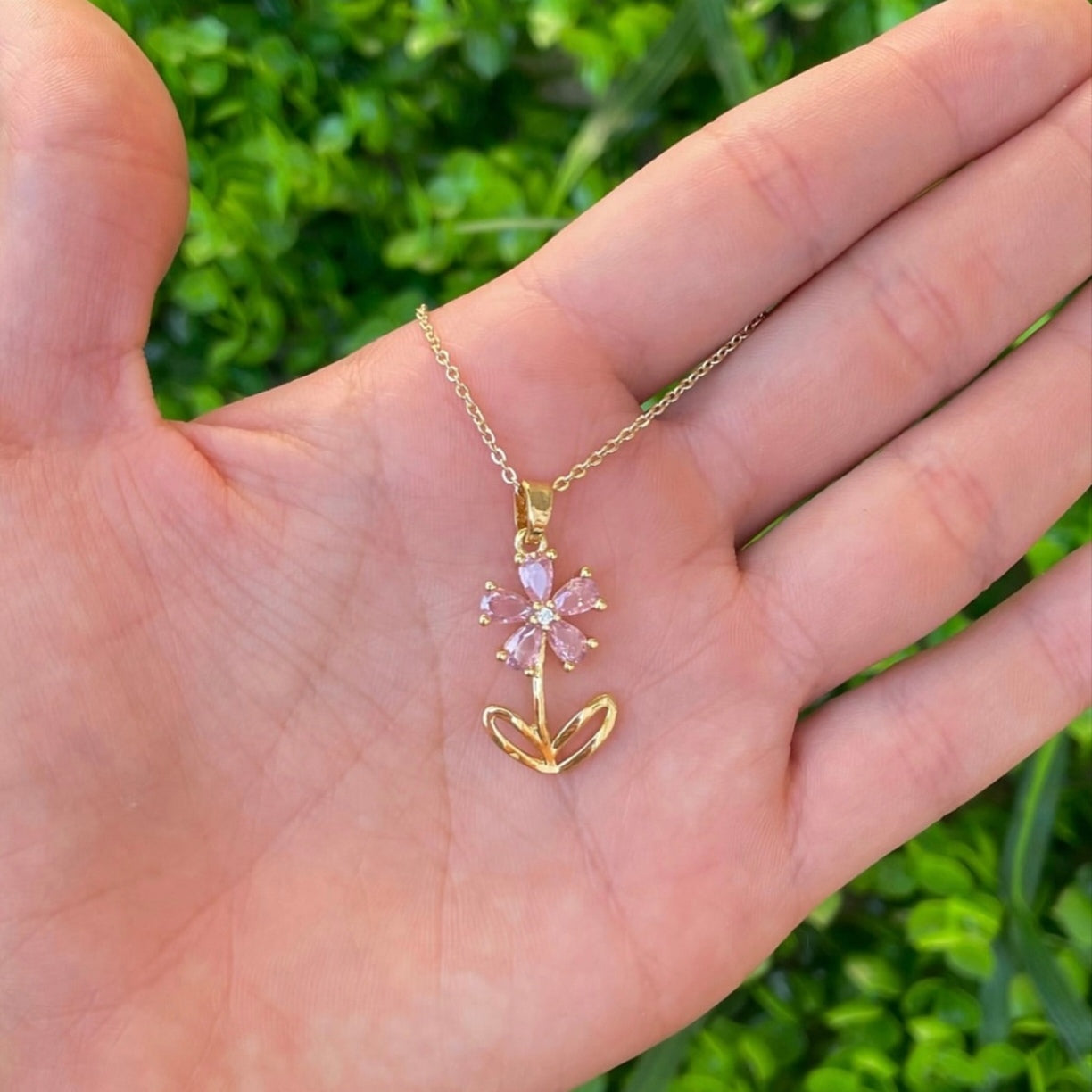 Gold necklace with a pink flower pendant held in a hand against a green background